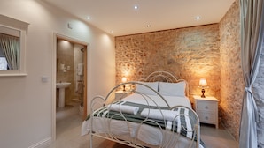 A bedroom with a stone wall, a bed with white linens, a nightstand with a lamp, and a bathroom with a sink and mirror. - HILLS FARM STAYS (Axminster)