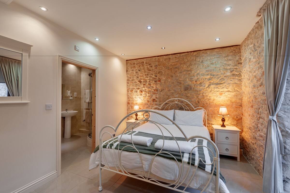 Un dormitorio con pared de piedra, una cama con ropa de cama blanca, una mesita de noche con lámpara, y un baño con lavamanos y espejo.