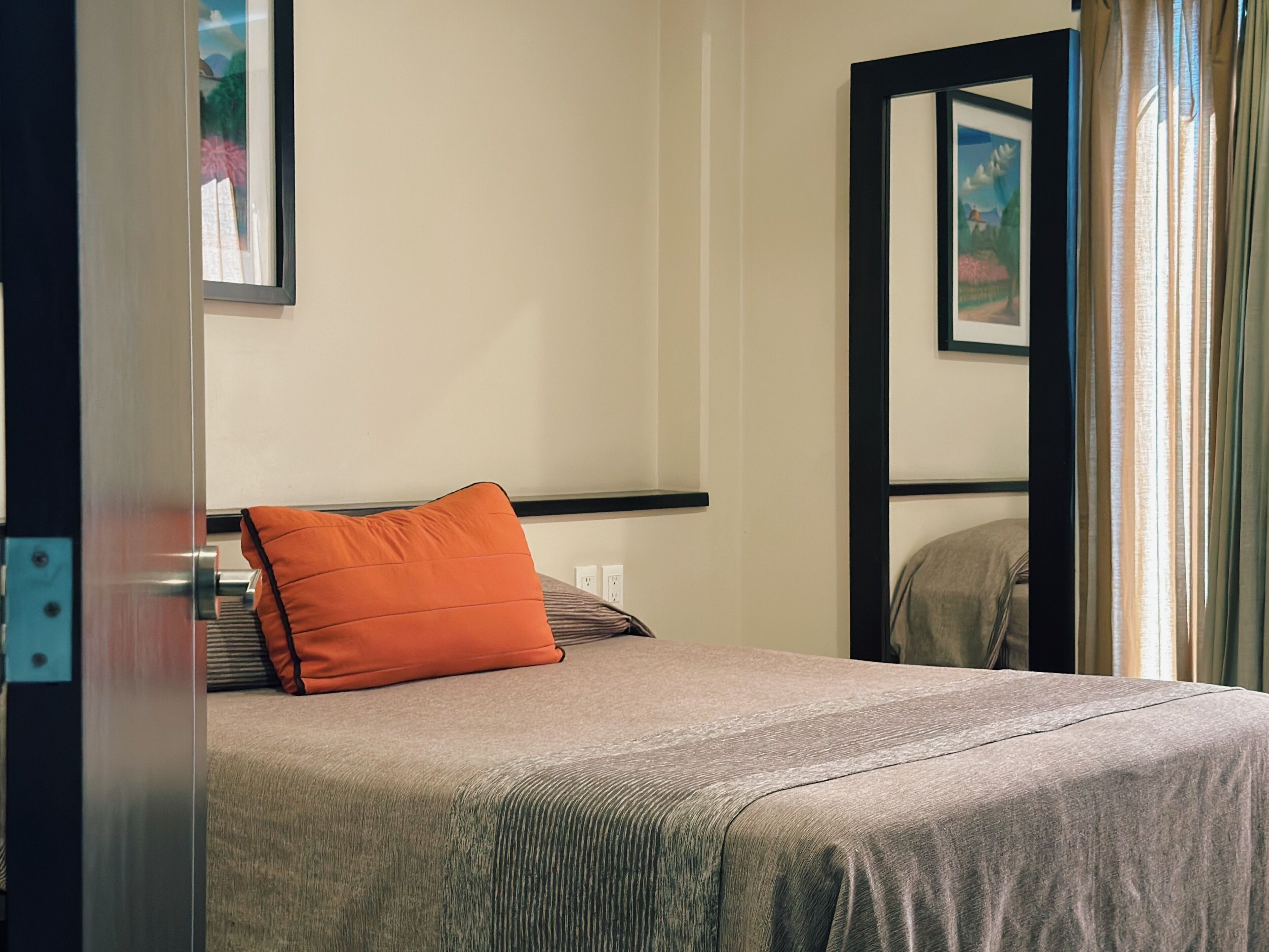 Een net opgemaakt bed met een grijze dekbedovertrek en een oranje kussen, een ingelijste foto aan de muur, en een raam met gordijnen.