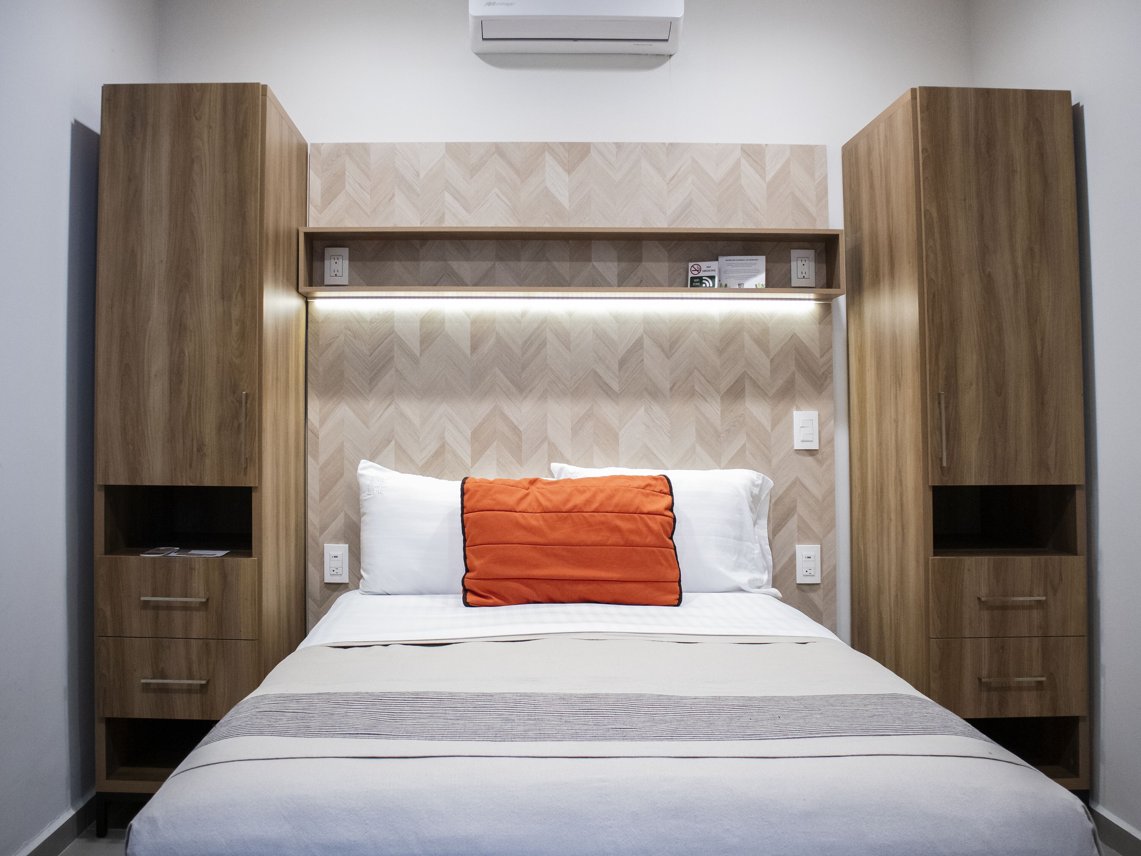 A hotel room with a bed, two wooden wardrobes, a built-in shelf, and a wall-mounted air conditioner.