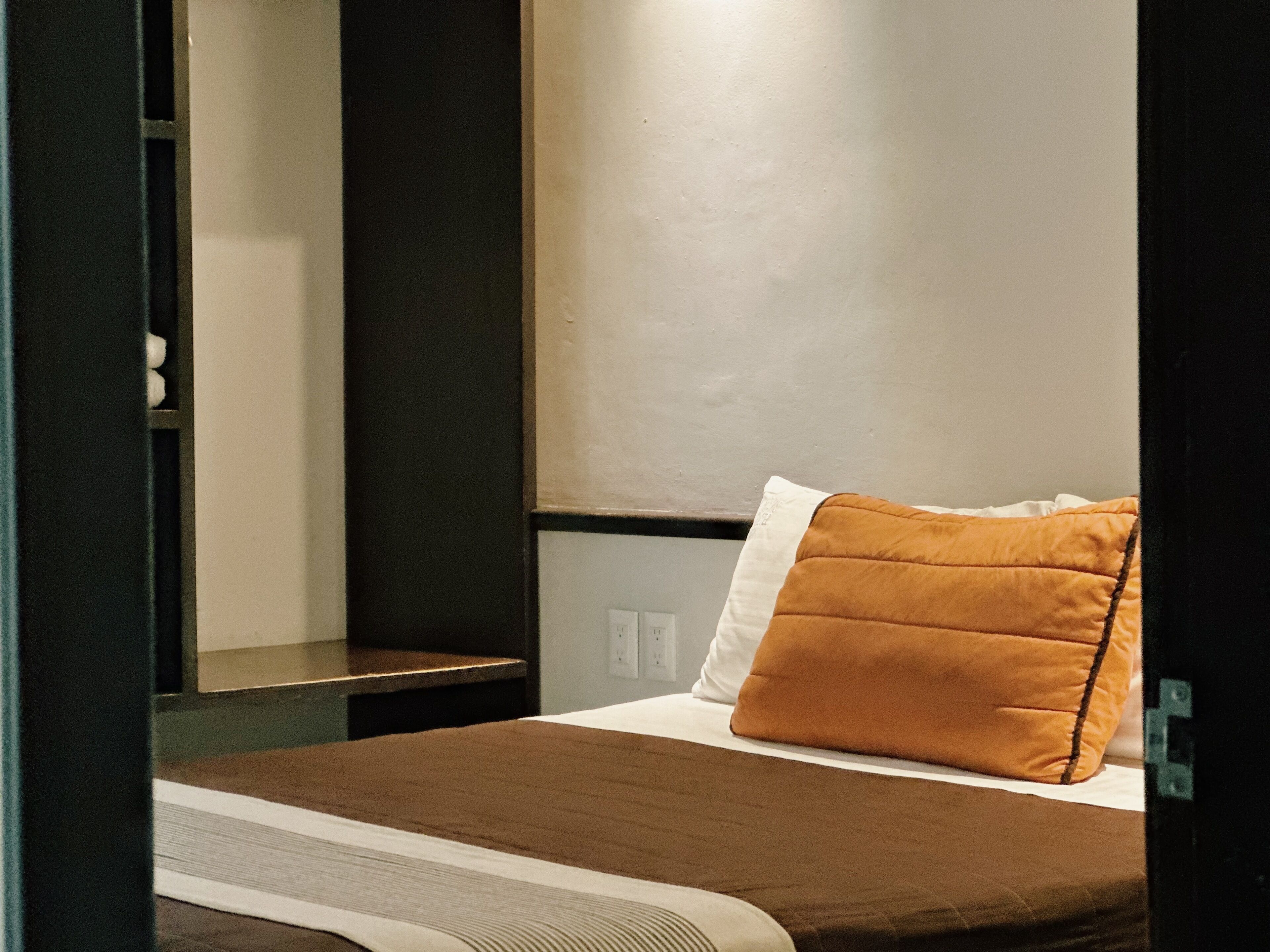 A neatly made bed with a brown blanket and a single orange pillow.