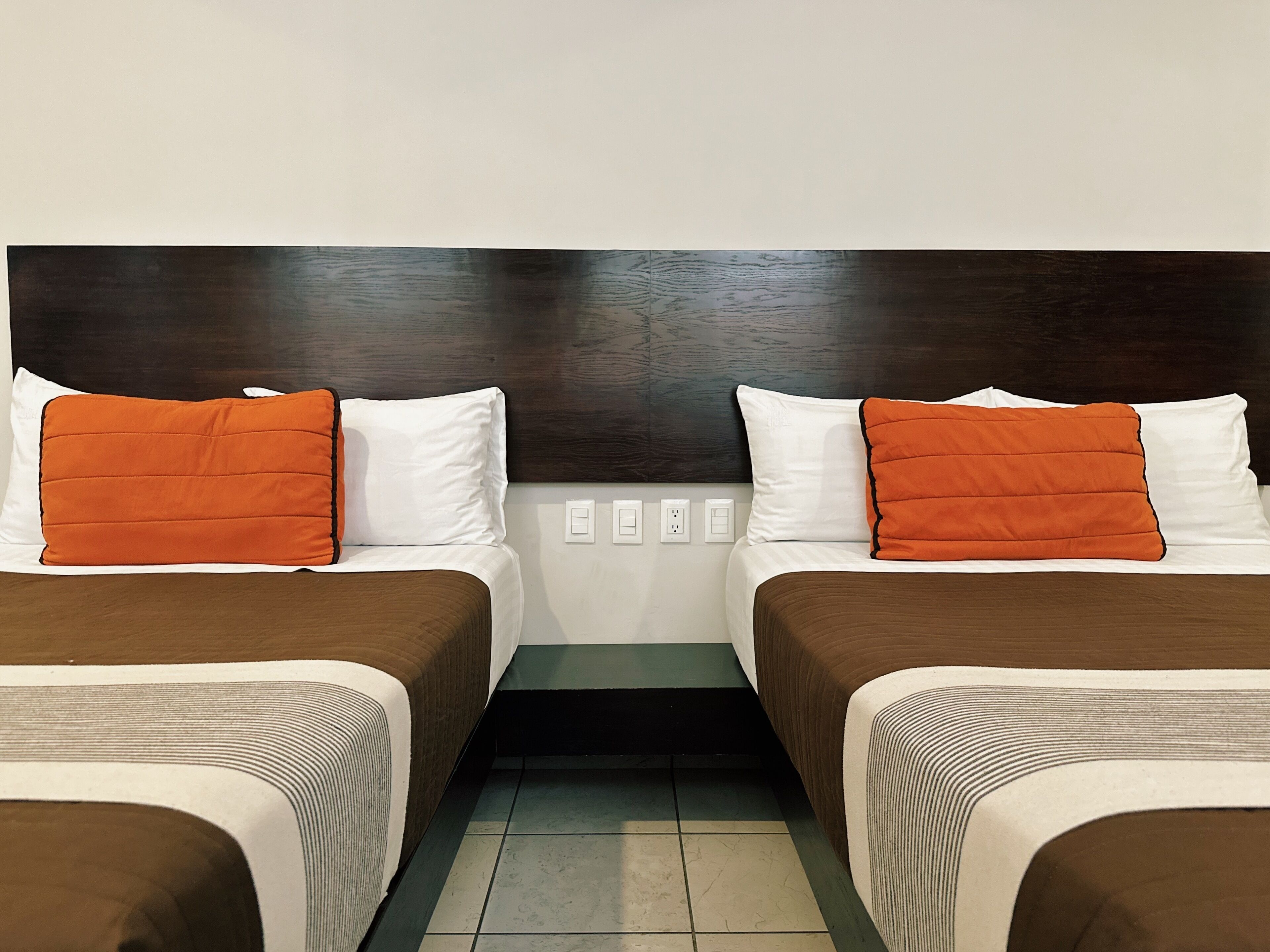 Habitación de hotel con dos camas, cada una con cabecero marrón y ropa de cama blanca. Hay dos mesitas de noche blancas con una lámpara pequeña en cada una.
