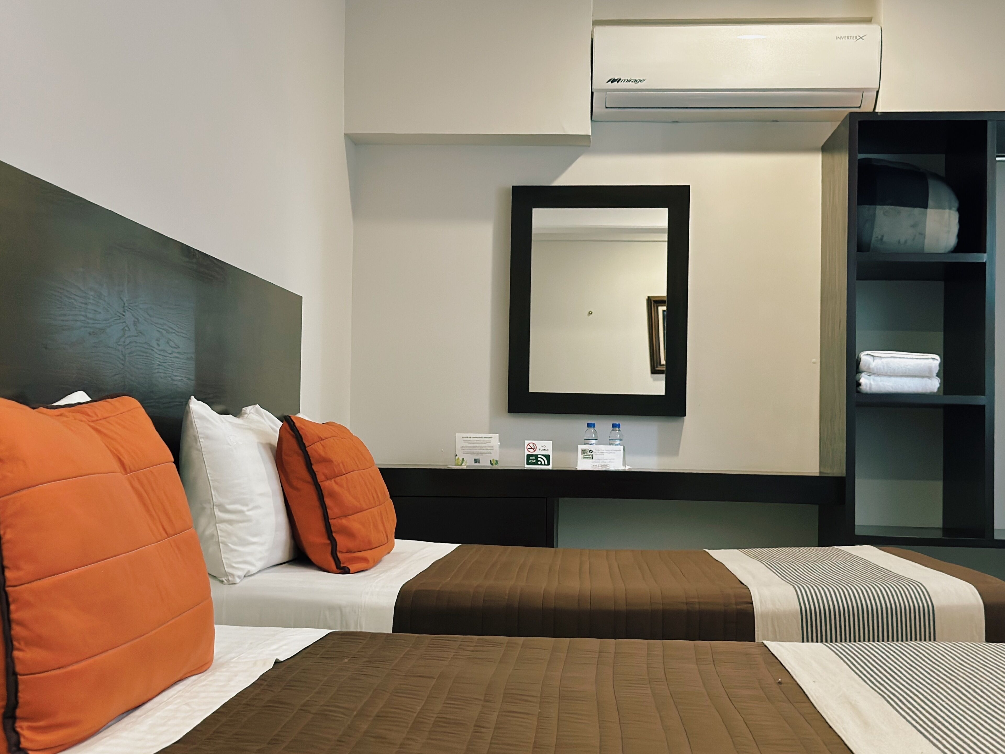 A hotel room with two beds, a bedside table with a mirror, a shelf with towels, and an air conditioning unit.