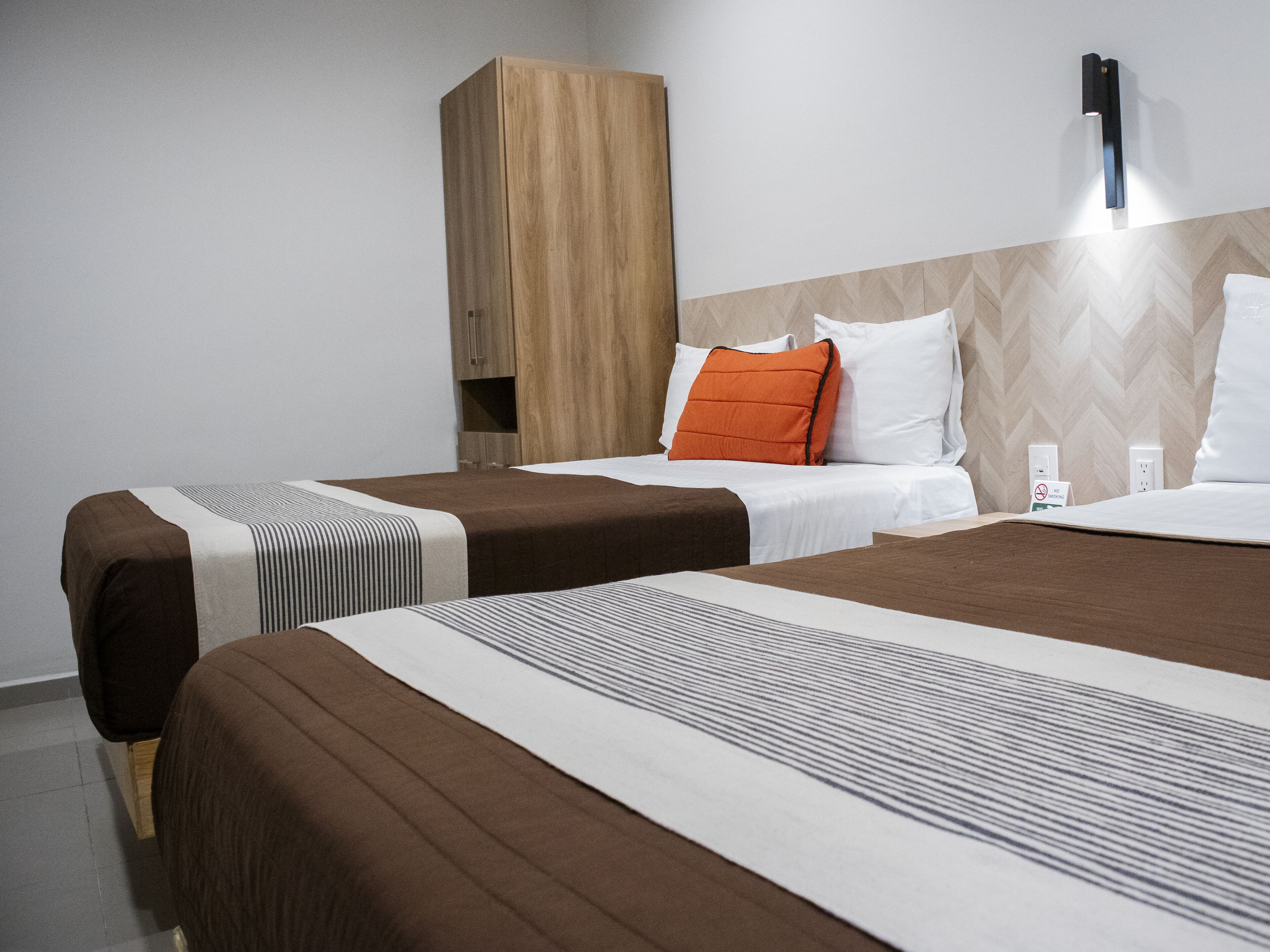 A hotel room with two beds, each with a brown bedspread and white pillows. There is a wooden wardrobe and a wall-mounted light fixture.