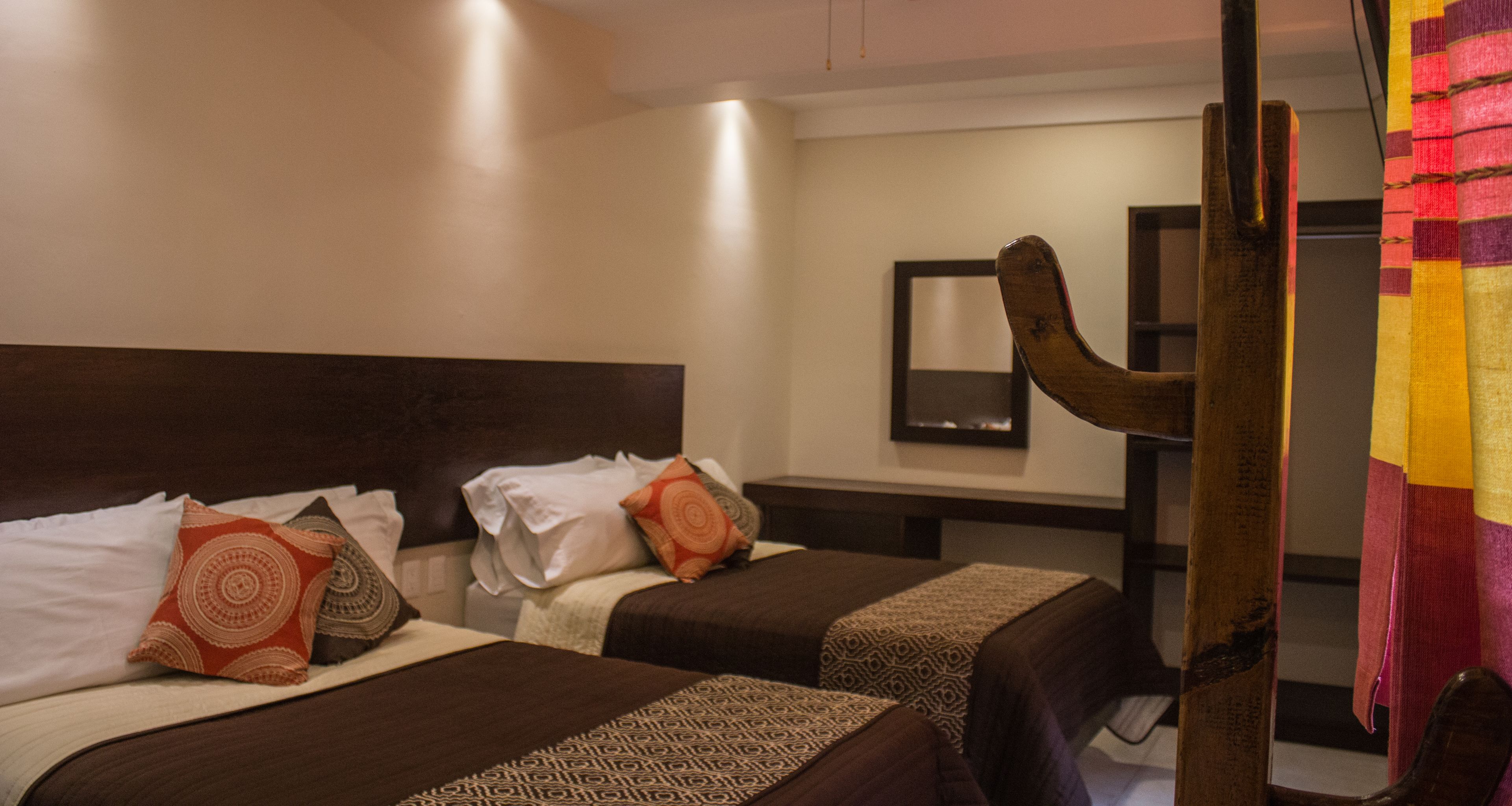 A hotel room with two beds, each with patterned bedding and pillows. There is a wooden headboard, a mirror on the wall, and a curtain with a colorful pattern.