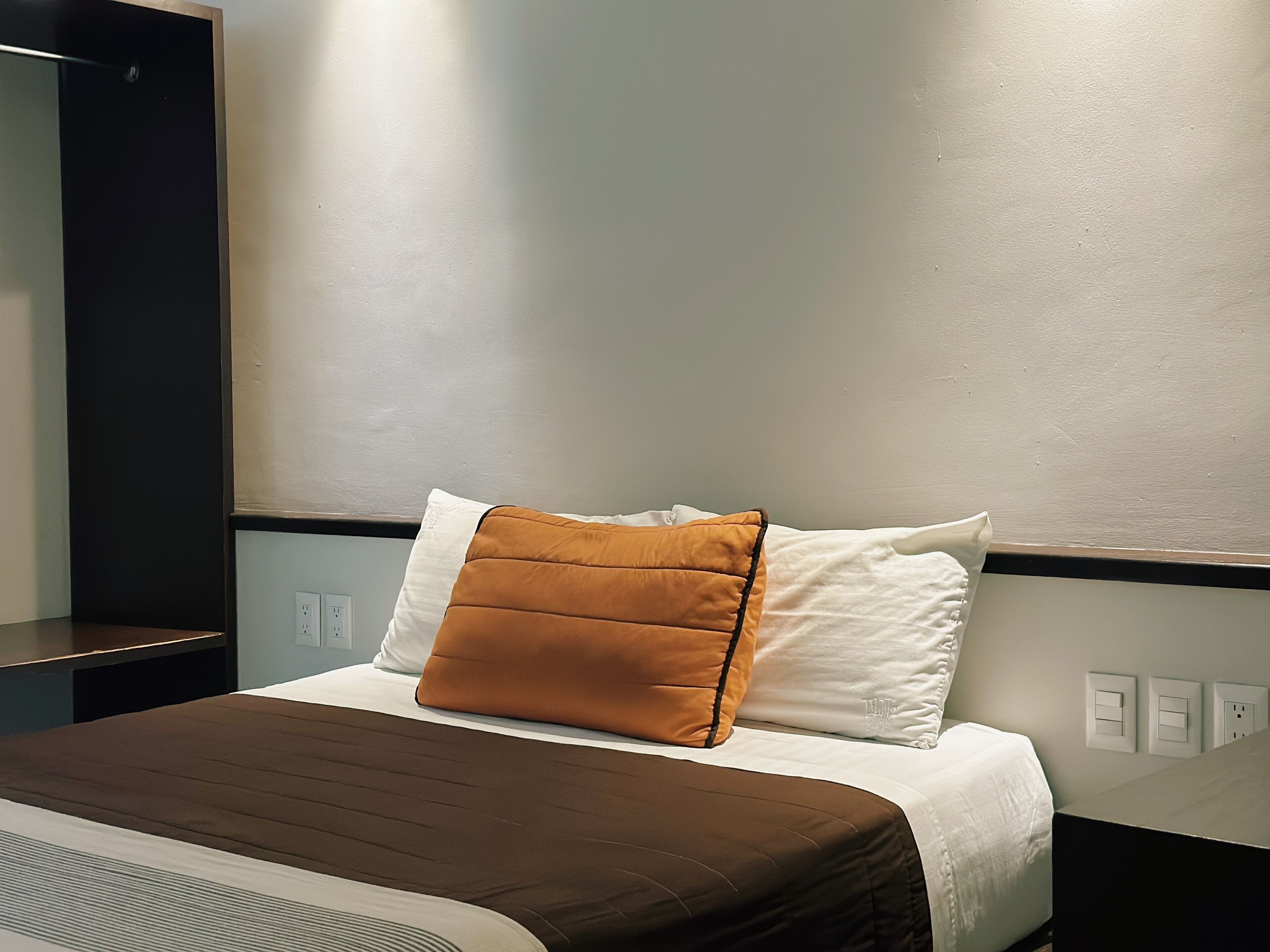 A hotel room with a neatly made bed, a brown bedspread, white pillows, and a brown accent pillow.