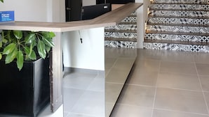 Un mostrador de recepción con un cartel que dice "MAYIS HOTEL" y un letrero de "SOLD PERSONAL" sobre una puerta. Una escalera con baldosas estampadas conduce a un piso superior.
