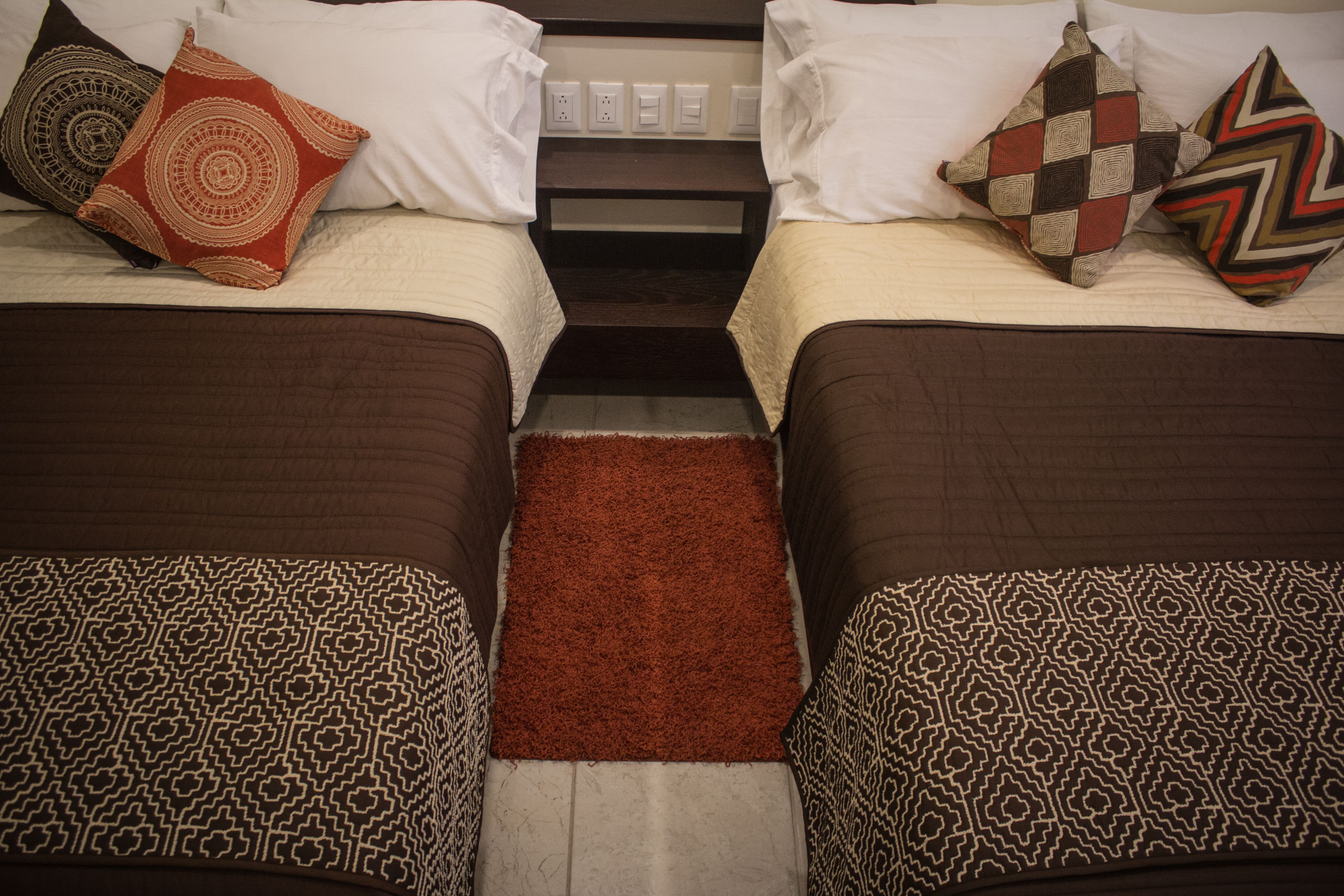 Une chambre d’hôtel avec deux lits, chacun agrémenté de draps et d’oreillers à motifs, un petit tapis et une table de chevet avec une lampe.