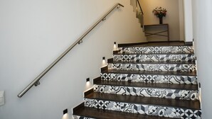 Una escalera con baldosas estampadas y barandilla de metal, que conduce a un rellano con una pequeña mesa y un jarrón de flores.
