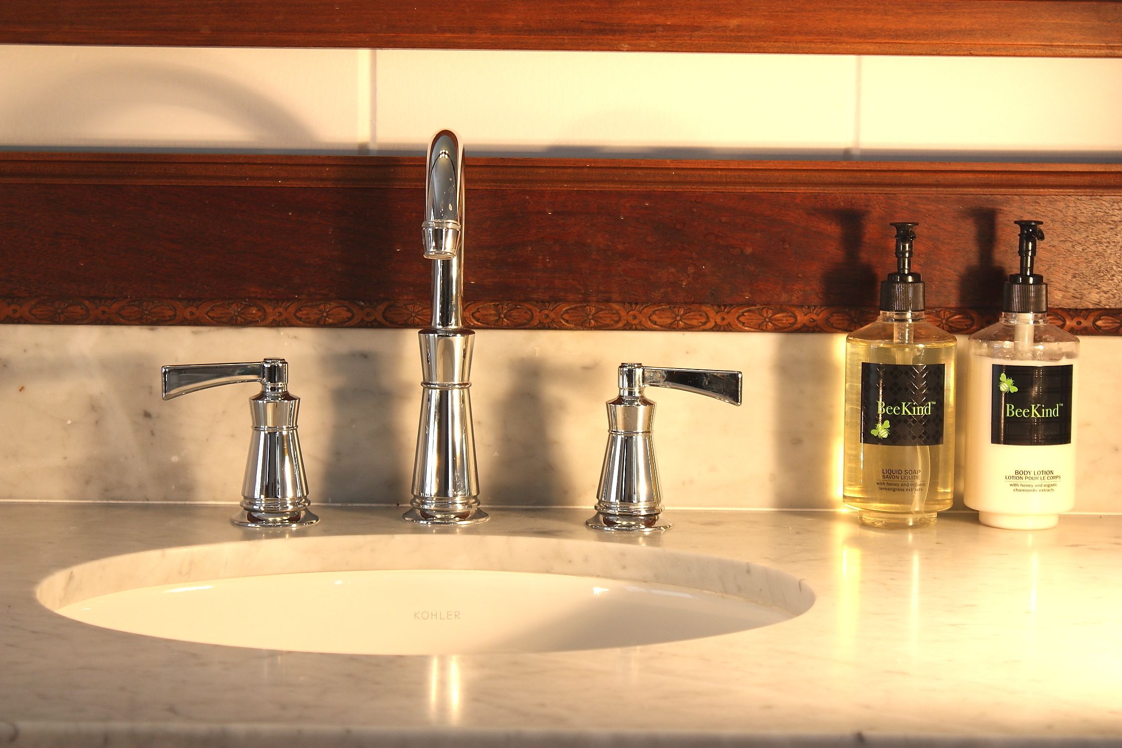 A bathroom sink with three faucets, two bottles of BeeKind body lotion, and a marble countertop.