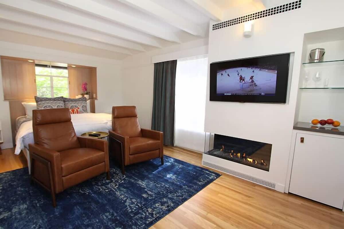 A modern living room with a fireplace, a TV mounted on the wall, a bed with a window view, and a seating area with two chairs.