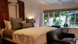 A bedroom with a bed, a leather chair, a bedside table, and a window with a view of greenery. - The Vagabond's House (Carmel)
