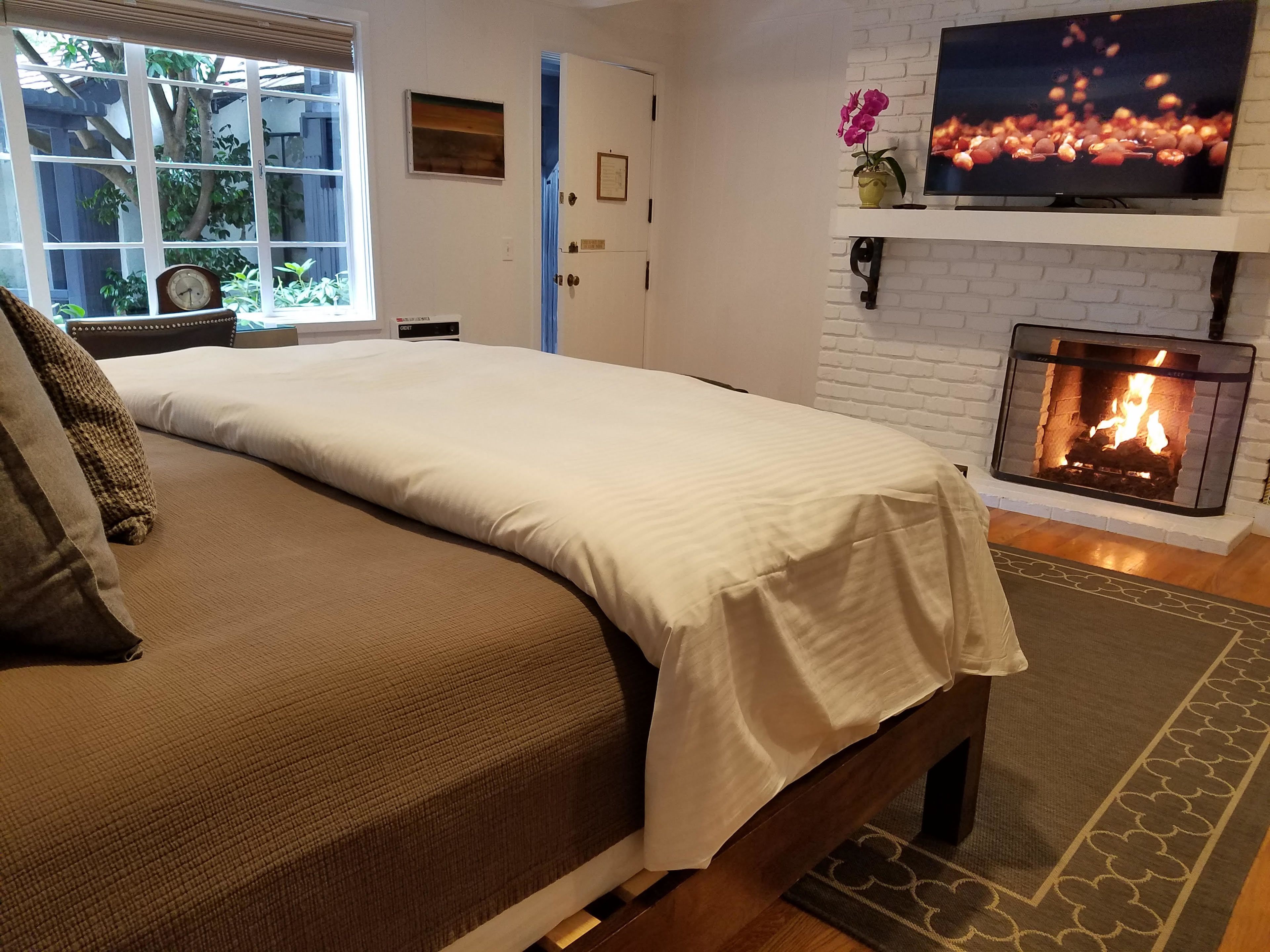 A bedroom with a bed, a fireplace, a TV, and a window with a view of greenery.