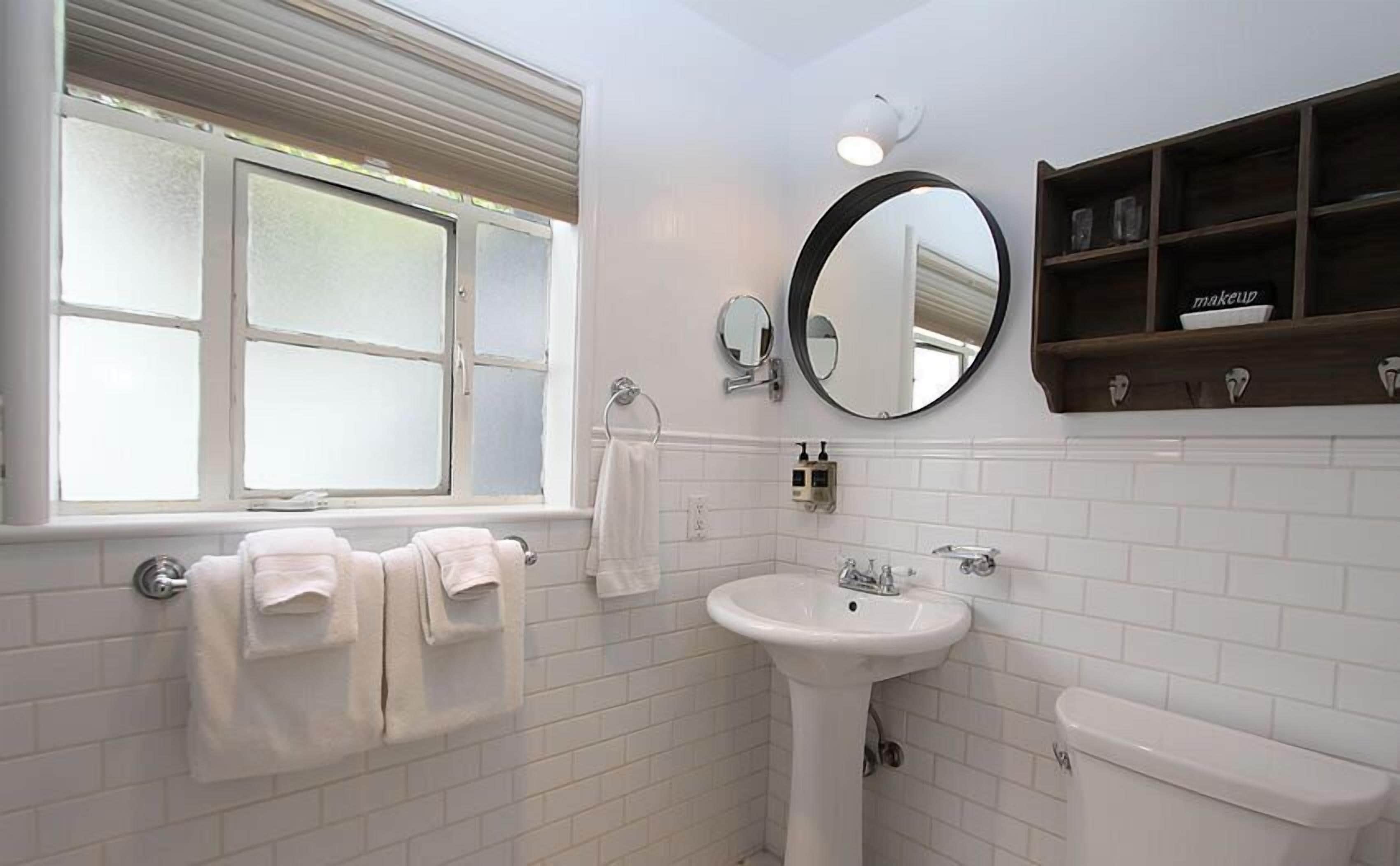 Ein Badezimmer mit weißen Fliesen, einem Fenster mit Jalousien, einem Waschbecken, einer Toilette und einem Spiegel über dem Waschbecken.
