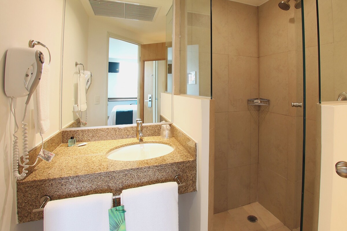 Une salle de bain dotée d’un comptoir en granit, d’un lavabo, d’un miroir, d’un sèche-cheveux et d’un espace douche.
