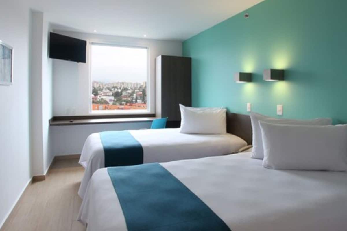Ein Hotelzimmer mit zwei Betten, einem Fenster mit Blick auf die Stadt, einem Flachbildfernseher und Wandleuchten.