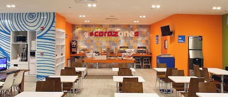 A cafeteria with tables and chairs, a counter with food items, and a patterned wall with text.