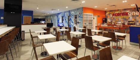 Una cafetería amplia con varias mesas y sillas, una barra con un menú y un pared con diseño en el fondo.
