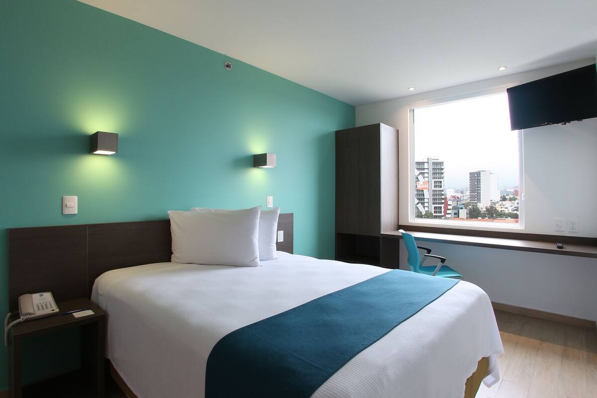 Ein Hotelzimmer mit einem Bett, einem Nachttisch mit Telefon, einem Fenster mit Blick auf die Stadt und einem an der Wand befestigten Fernseher.