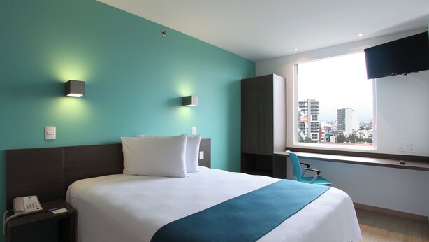 Een hotelkamer met een bed, nachtkastje, stoel, raam met uitzicht over de stad en een aan de muur bevestigde televisie.