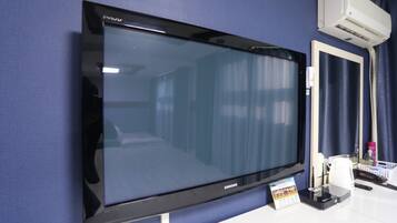 A flat-screen TV mounted on a blue wall, a white air conditioning unit above it, and a small table with a remote control and a card.