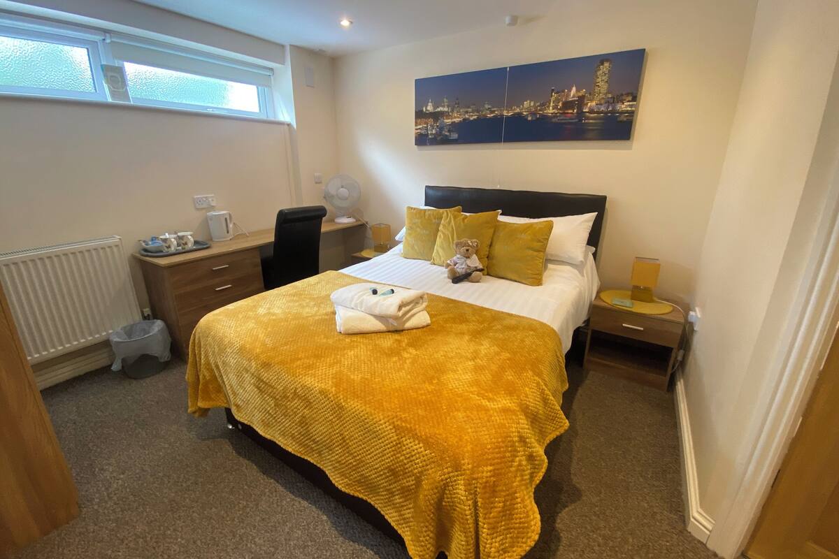 Une chambre d’hôtel avec un lit, un bureau, une chaise, un radiateur, une fenêtre et un tableau représentant un paysage urbain accroché au mur.