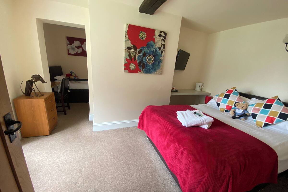 Une chambre avec un couvre-lit rouge, un bureau avec une chaise, une petite table avec une bouilloire et un ours en peluche sur le lit.