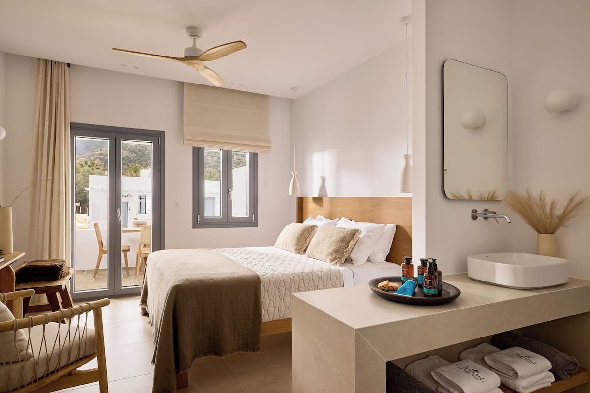 A hotel room with a bed, a desk, a chair, a bathroom with a sink, and a view of the outside.
