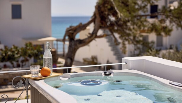 Un jacuzzi con una botella y un vaso sobre una bandeja de madera, con vista a una playa y un edificio blanco.