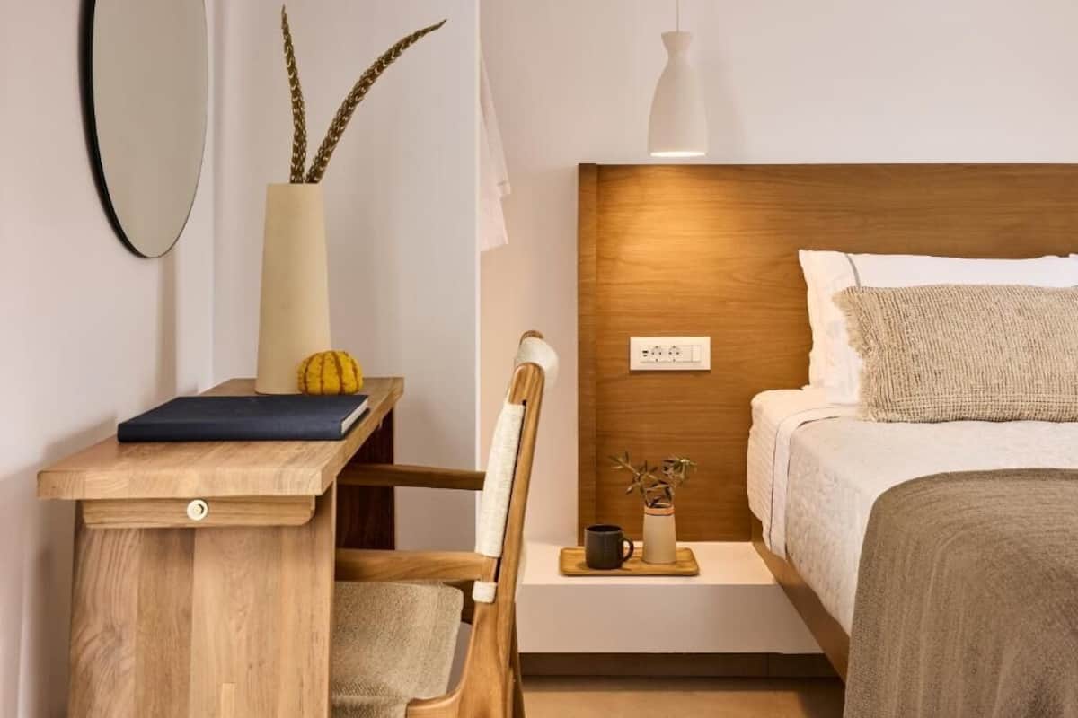 A bedroom with a wooden headboard, a bedside table with a book, a vase with dried plants, and a small nightstand with a lamp.