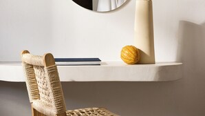 Una habitación moderna con un espejo redondo, una silla de madera, un estante blanco con un jarrón y un libro.