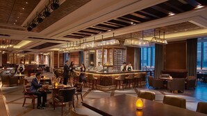 A spacious hotel lobby with a bar area, multiple seating arrangements, and a view of the cityscape through large windows.