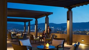 An outdoor seating area with wicker furniture and a table with a candle, overlooking a cityscape at dusk.
