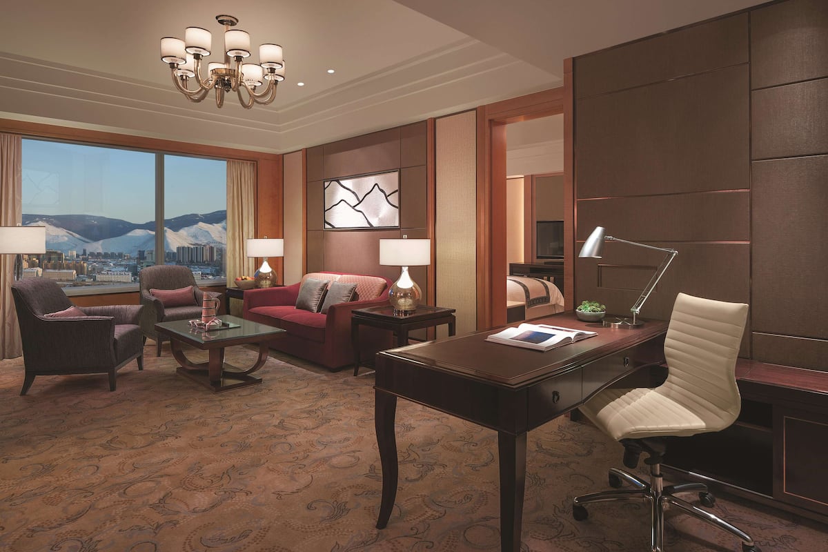 Hotelkamer met groot raam met uitzicht op de stad, een kroonluchter, een bank, een bureau en een stoel.