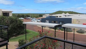 A view from a balcony overlooking a parking area with several cars, a building with a flat roof, and a hilly landscape in the distance. - Margaret River Beach Apartments (Gnarabup)
