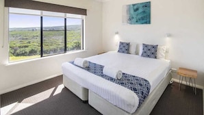 A bedroom with a large window, a bed with white and blue bedding, a small wooden bench, and a painting on the wall. - Margaret River Beach Apartments (Gnarabup)