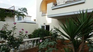 A building with a white facade, a balcony with metal railings, and a small garden with pink flowers and green plants. - Alexandra Hotel (Chania)