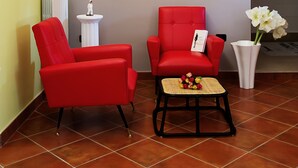 Une pièce avec une chaise rouge, une petite table ornée de fleurs, un vase blanc et un luminaire fixé au mur.