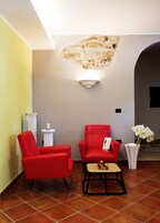 Una habitación con una silla roja, una mesita con flores, un jarrón blanco y una lámpara fijada a la pared.