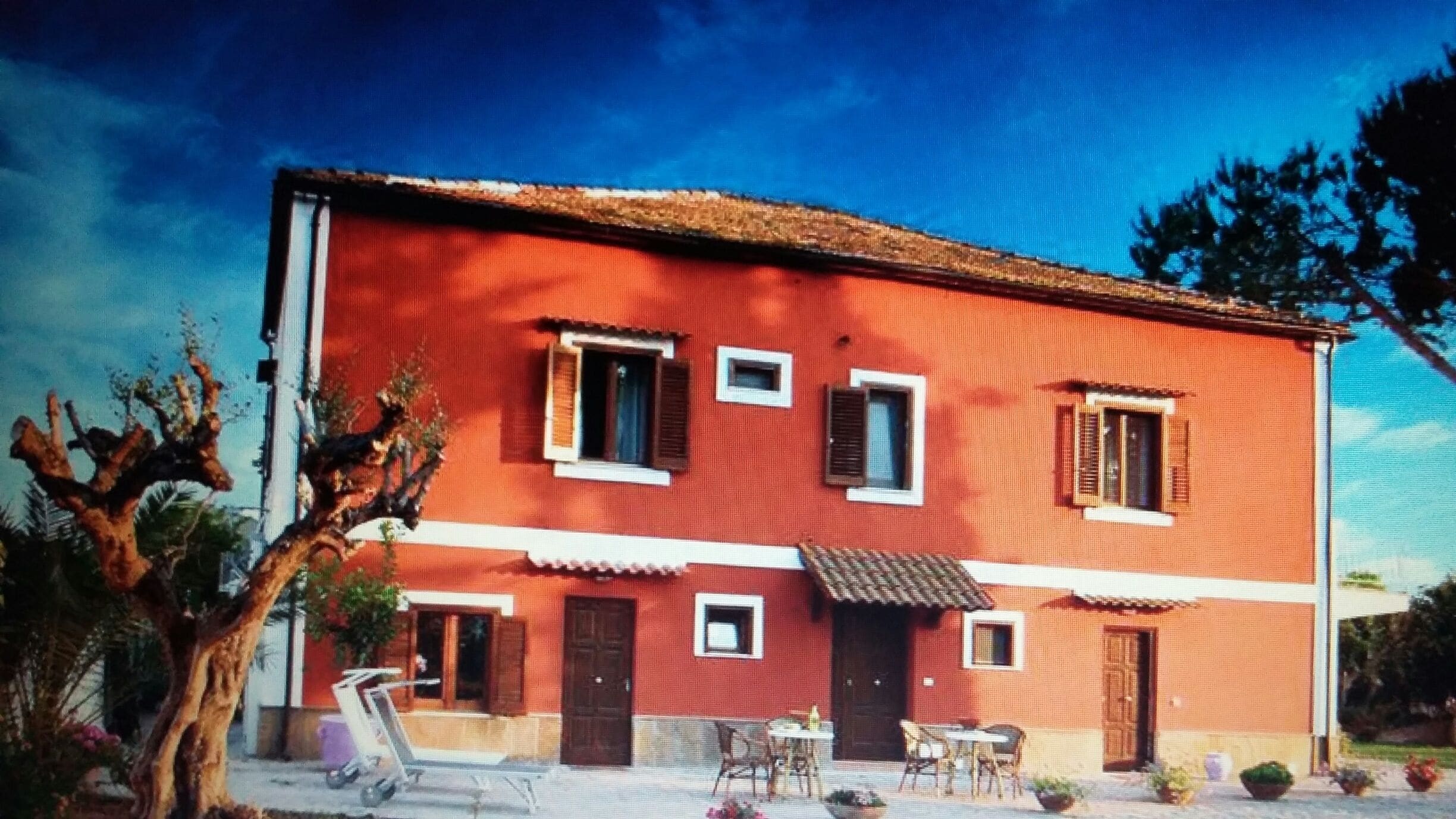 A red building with two windows and a door, a small table and chairs outside, and a tree in the foreground.