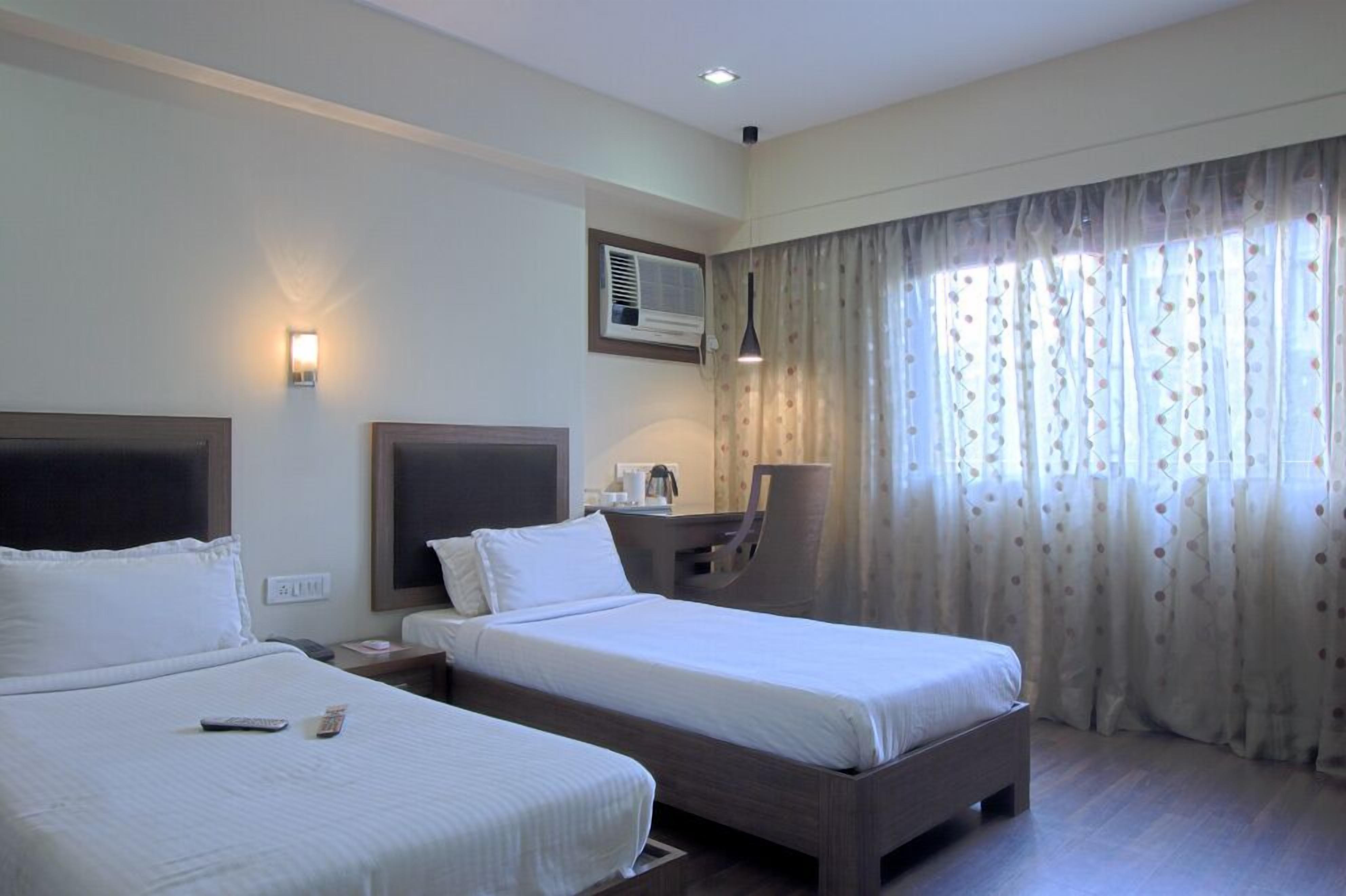Une chambre d’hôtel avec deux lits, un bureau, une chaise, une lampe et une fenêtre avec des rideaux.