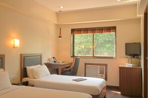 A hotel room with two beds, a desk, a chair, a television, and a window with a view of greenery. - Hotel Amigo (Mumbai)