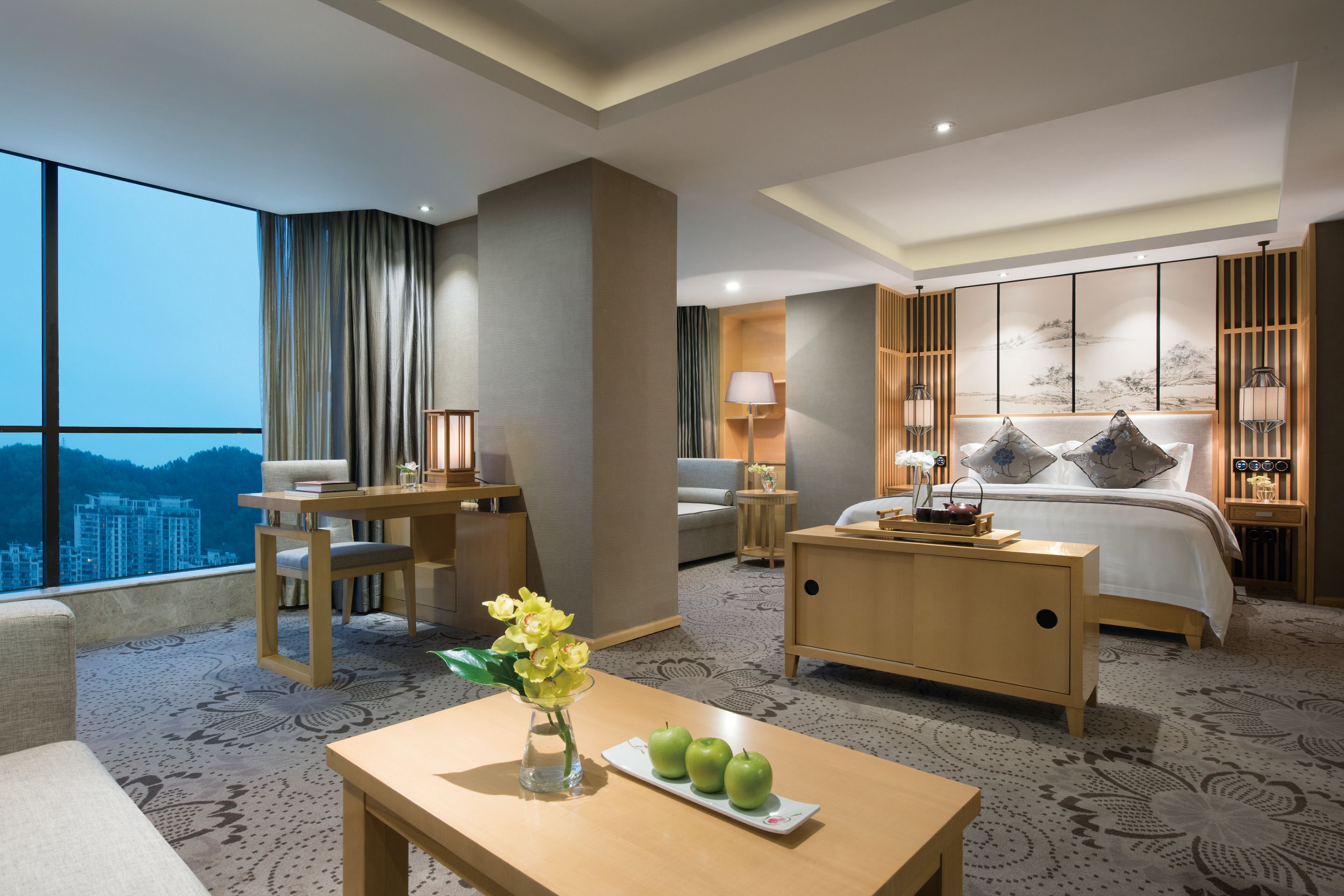 Habitación de hotel con un ventanal grande, una cama, un escritorio, una silla, una mesita de noche y una mesa con manzanas y flores.