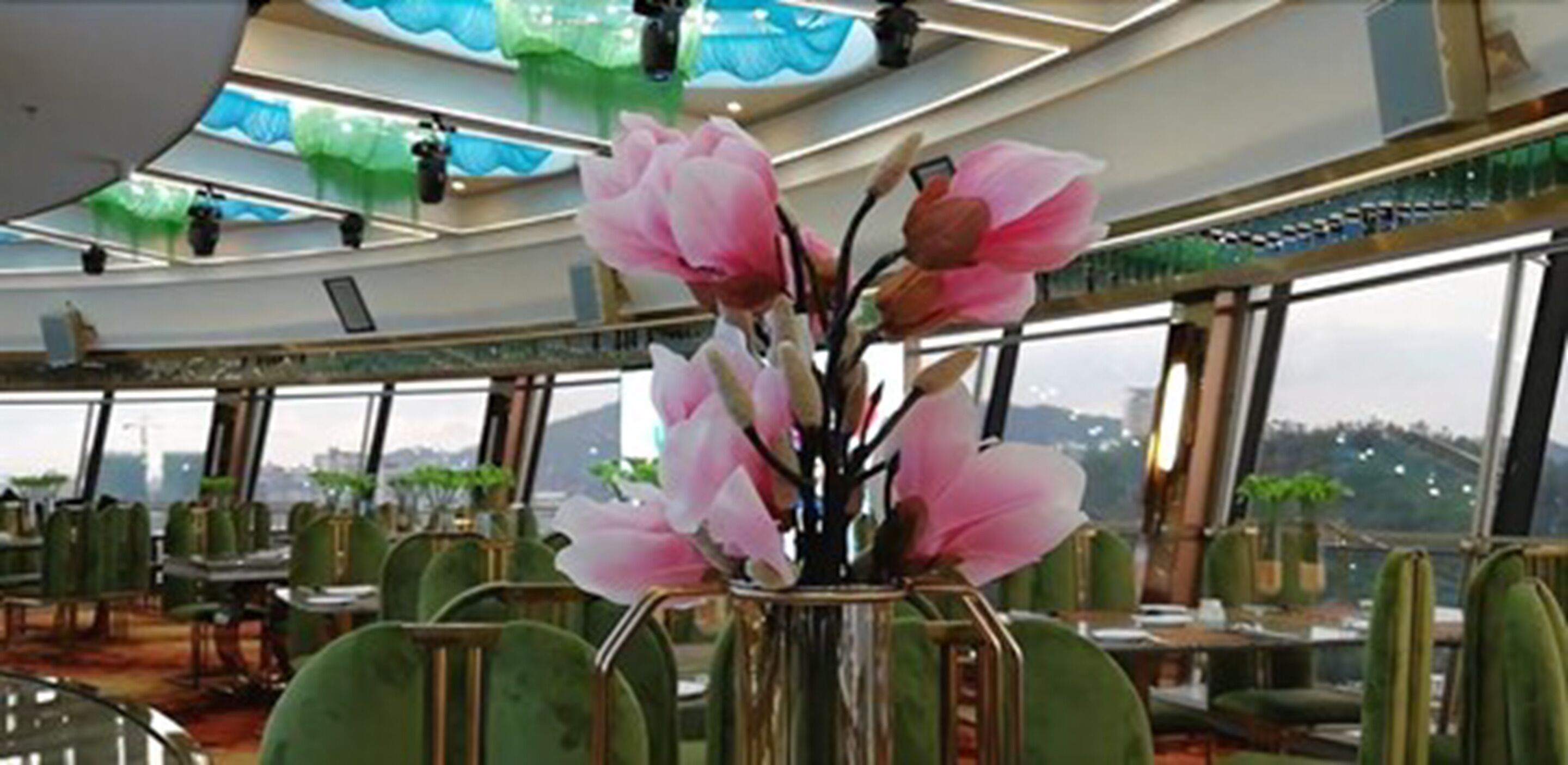 A restaurant with a large window view, a vase with pink flowers, and green chairs.