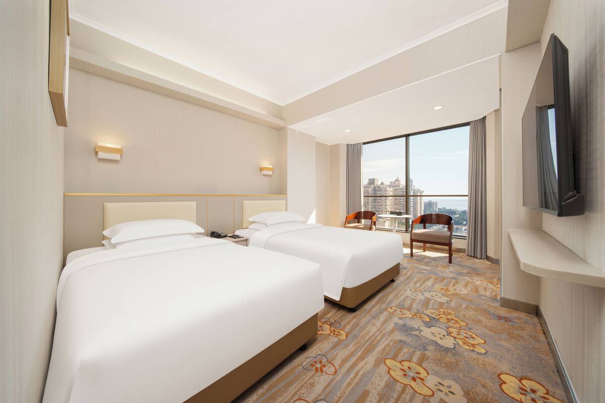 Ein Hotelzimmer mit zwei Betten, einem Fernseher, Blick auf die Stadt und einem großen Fenster.