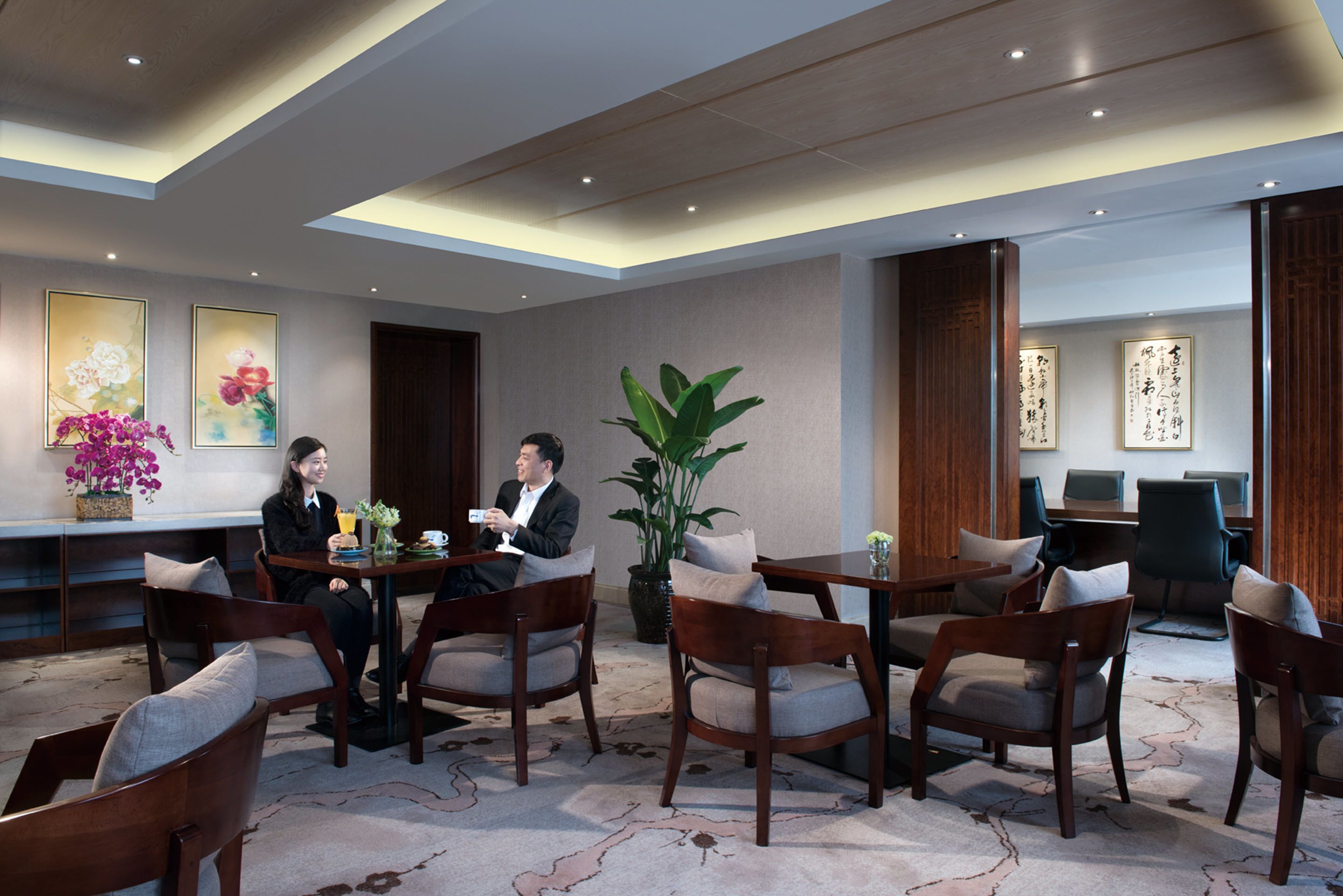 A modern hotel lobby with wooden furniture, a large plant, and artwork on the walls.