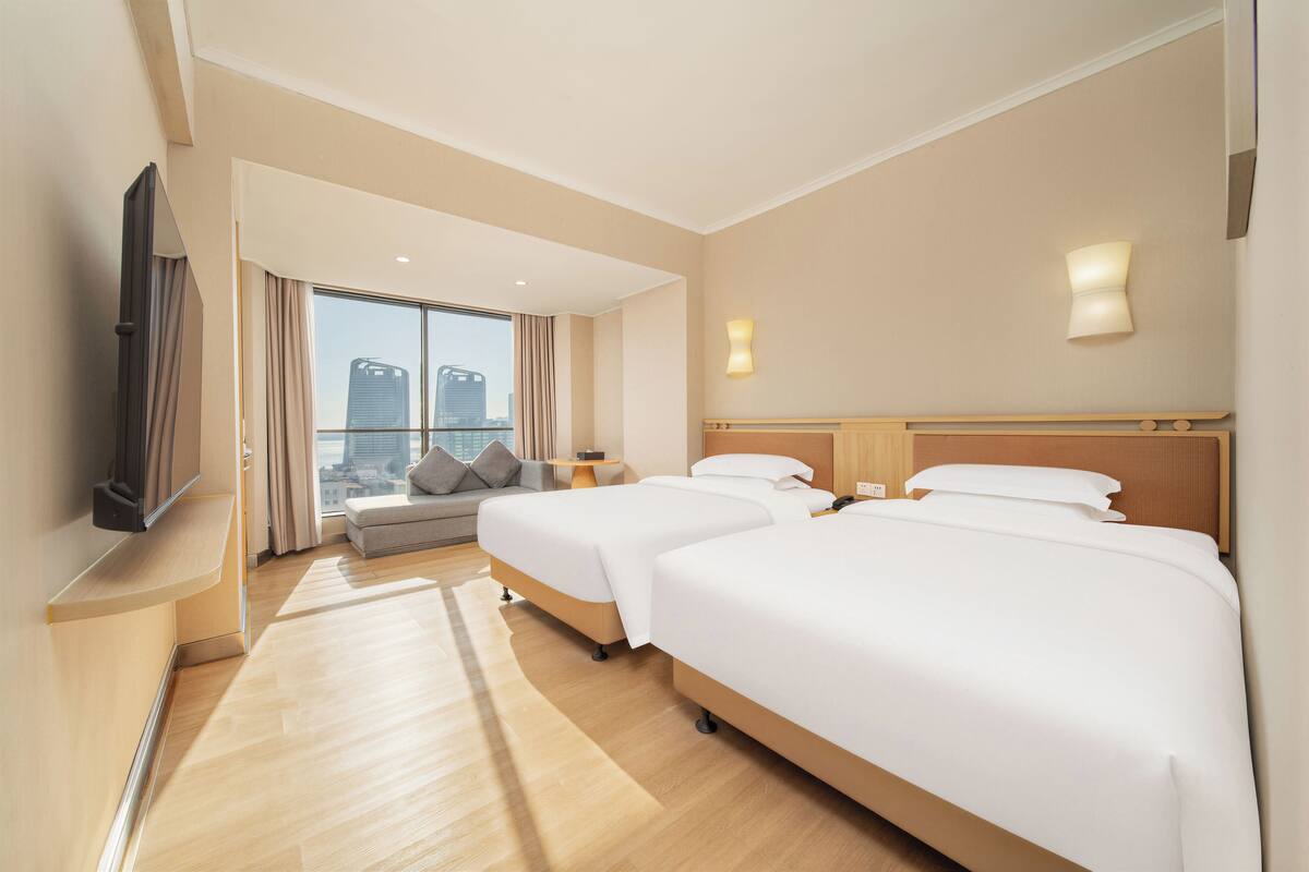 Ein Hotelzimmer mit zwei Betten, einem Schlafsofa, einem Fernseher und einem großen Fenster mit Blick auf die Stadt.