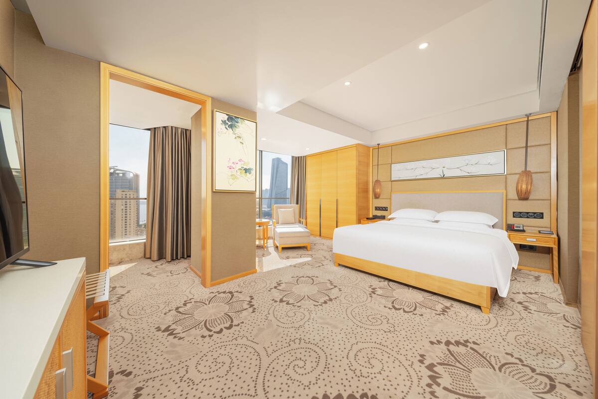 Ein Hotelzimmer mit einem großen Bett, einem Schreibtisch, einem Sessel, einem Fernseher und Blick auf die Stadt.