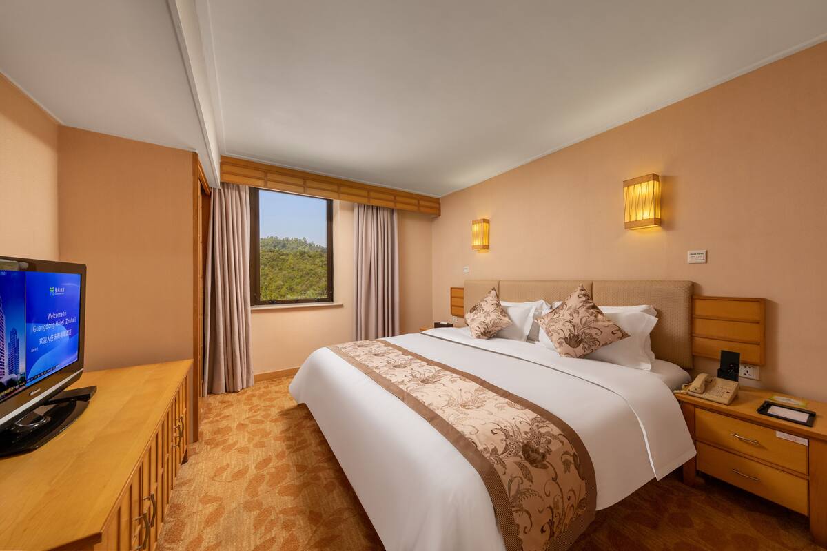 Ein Hotelzimmer mit einem großen Bett, einem Fernseher auf einem Holztisch, einem Nachttisch mit Telefon und einem Fenster mit Vorhängen.