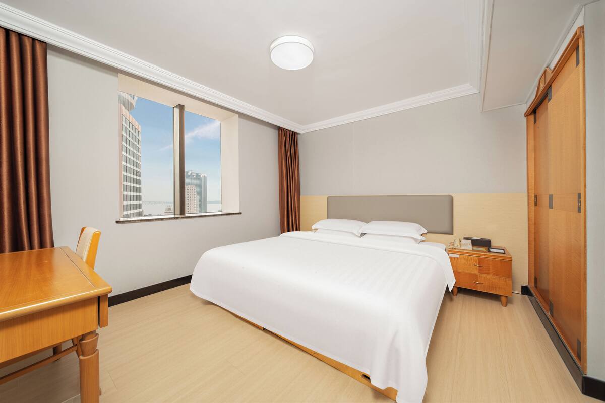 Ein Hotelzimmer mit Bett, Nachttisch aus Holz, Schreibtisch, Kleiderschrank und Fenster mit Blick auf die Stadt.